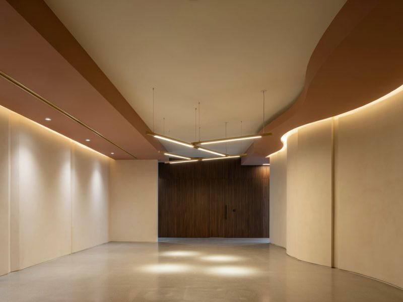A wide-angle view of a minimalist hallway featuring curved walls, warm LED strip lighting, and a dark wood door at the end of the corridor.